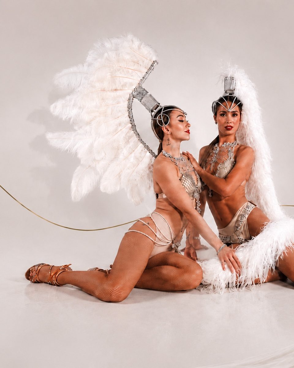 Two Refined showgirls in crystal costumes framed by large white feather wings