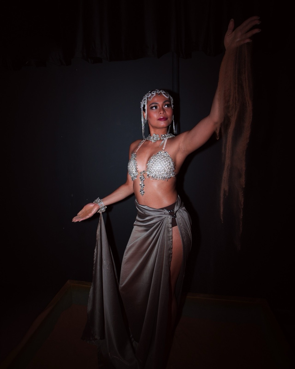 A dancer in a crystal-jewelled bra and chain headpiece holding a draped silk skirt against a dark backdrop