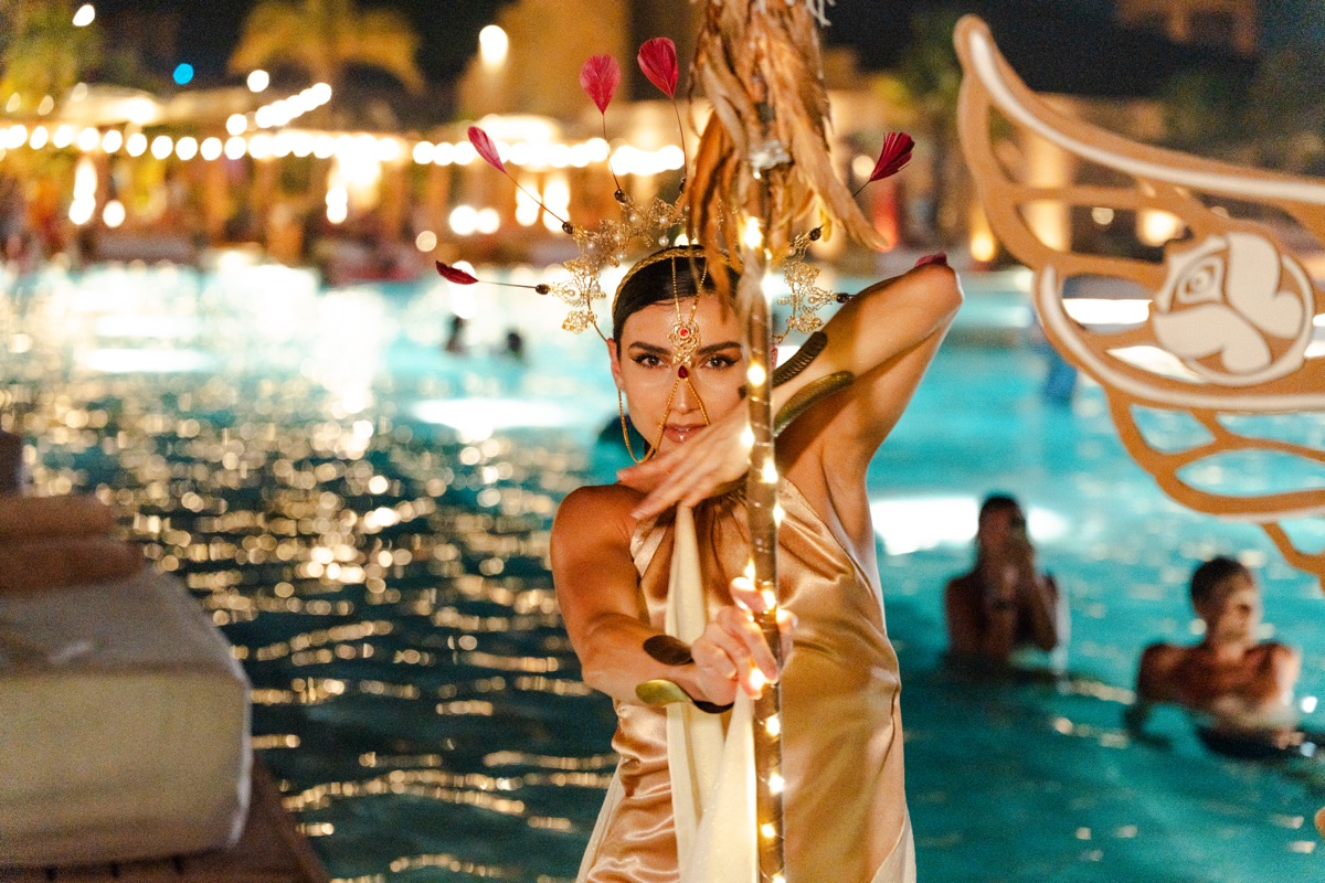 A dancer in gold with a red-feathered crown behind a light-wrapped pole at the pool's edge
