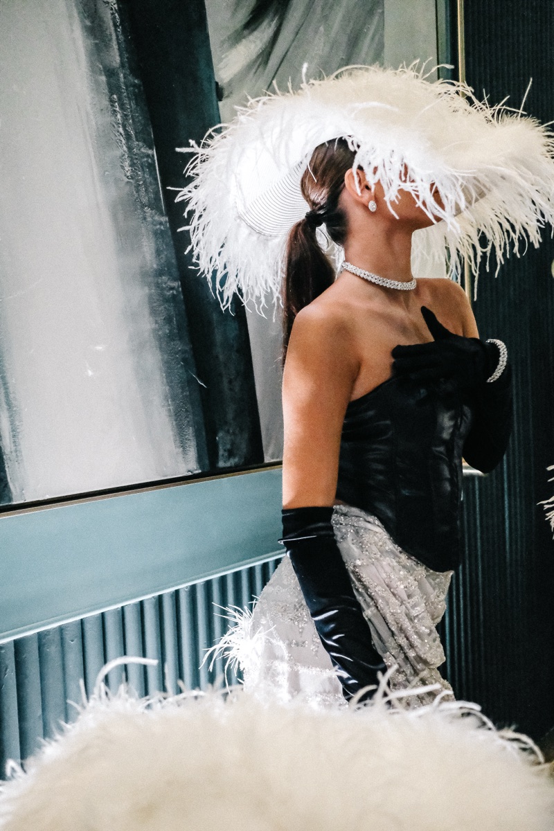 A performer in a white ostrich-feather hat, opera gloves and sequin skirt, seen from the side
