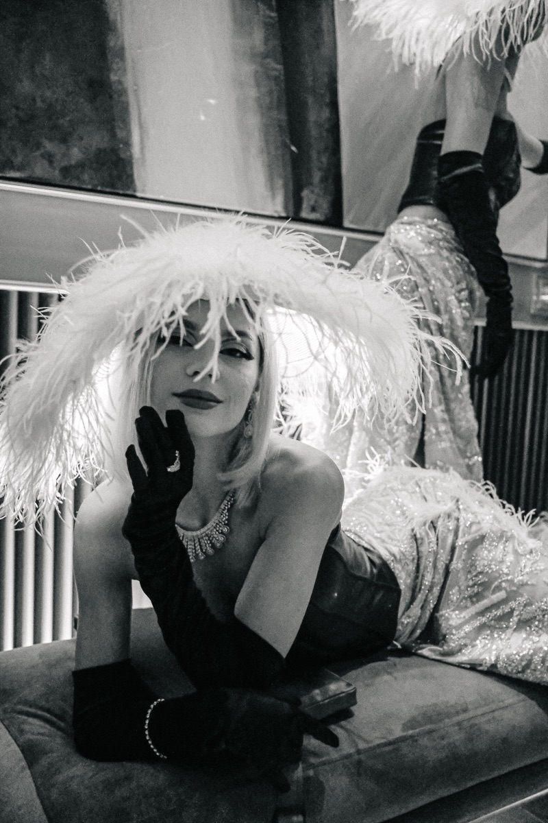 Two performers in feather hats and gowns reclining on a velvet bench, in black and white