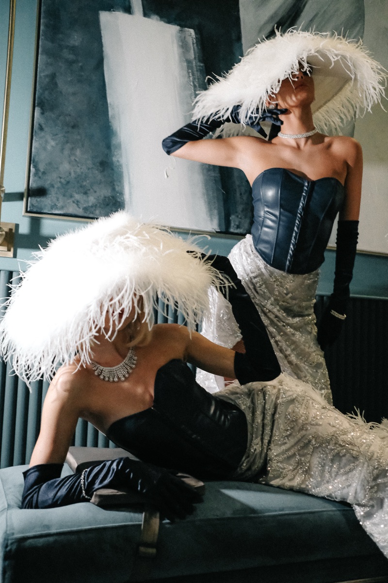 Two performers in white feather hats and corseted gowns, one reclining, one standing