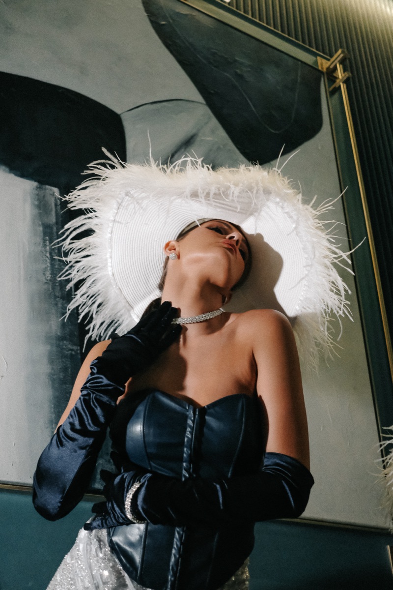 A low-angle portrait of a performer in a feather hat, corset and satin gloves