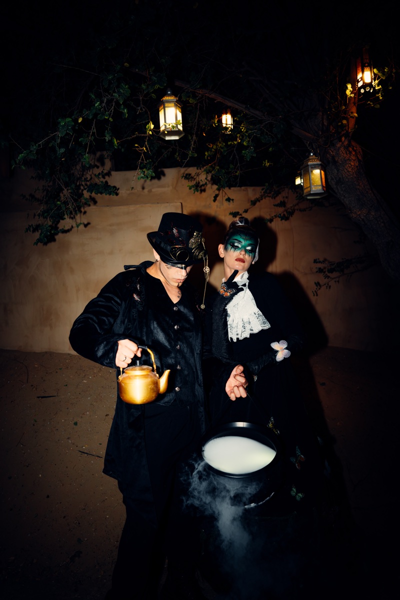Two masked Halloween performers pouring from a golden teapot into a smoking cauldron under lanterns