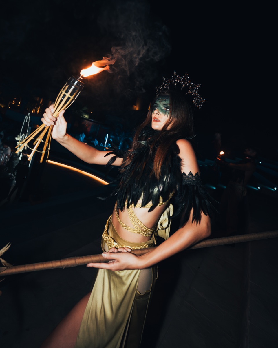 A crowned performer in a gold gown holding a flaming torch overhead at night