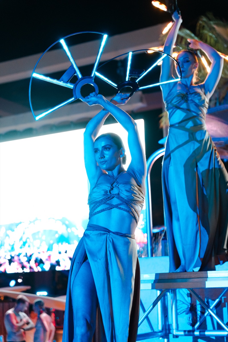 Two dancers holding arced LED fans overhead on lit platforms