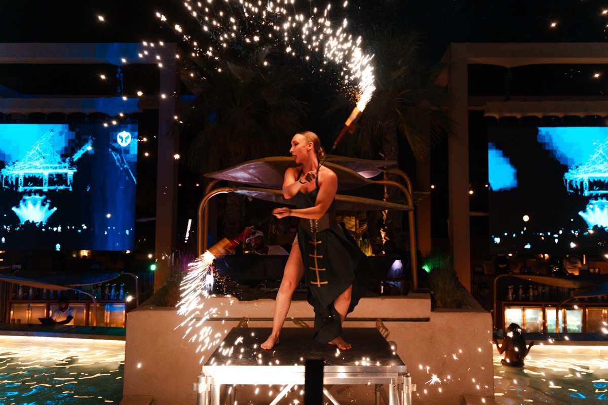 A performer seated on a poolside structure as a fountain of sparks erupts behind her