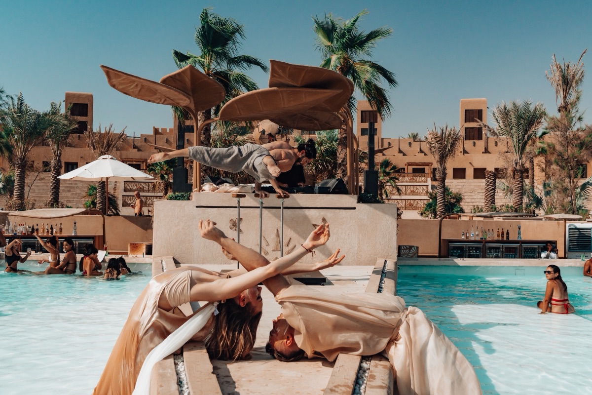 An acrobat balancing horizontally on a poolside structure while two dancers arch below in daylight