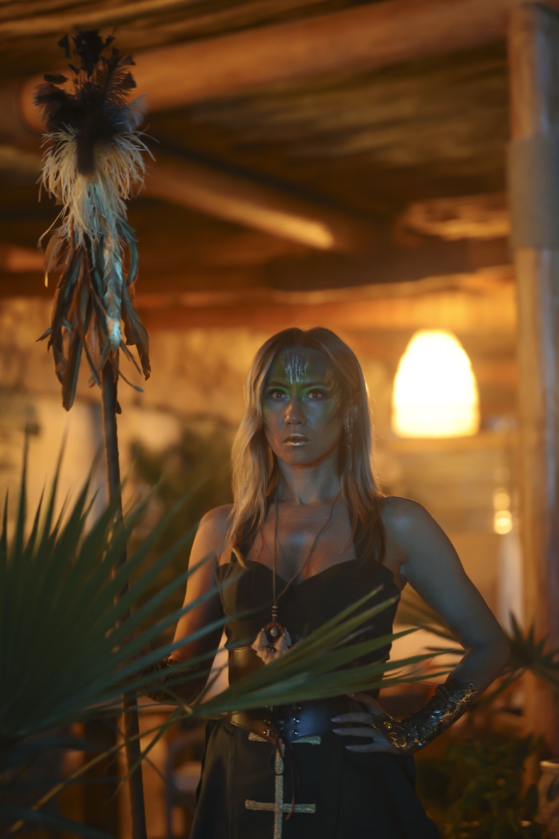 A performer with turquoise face paint holding a feathered staff under warm light