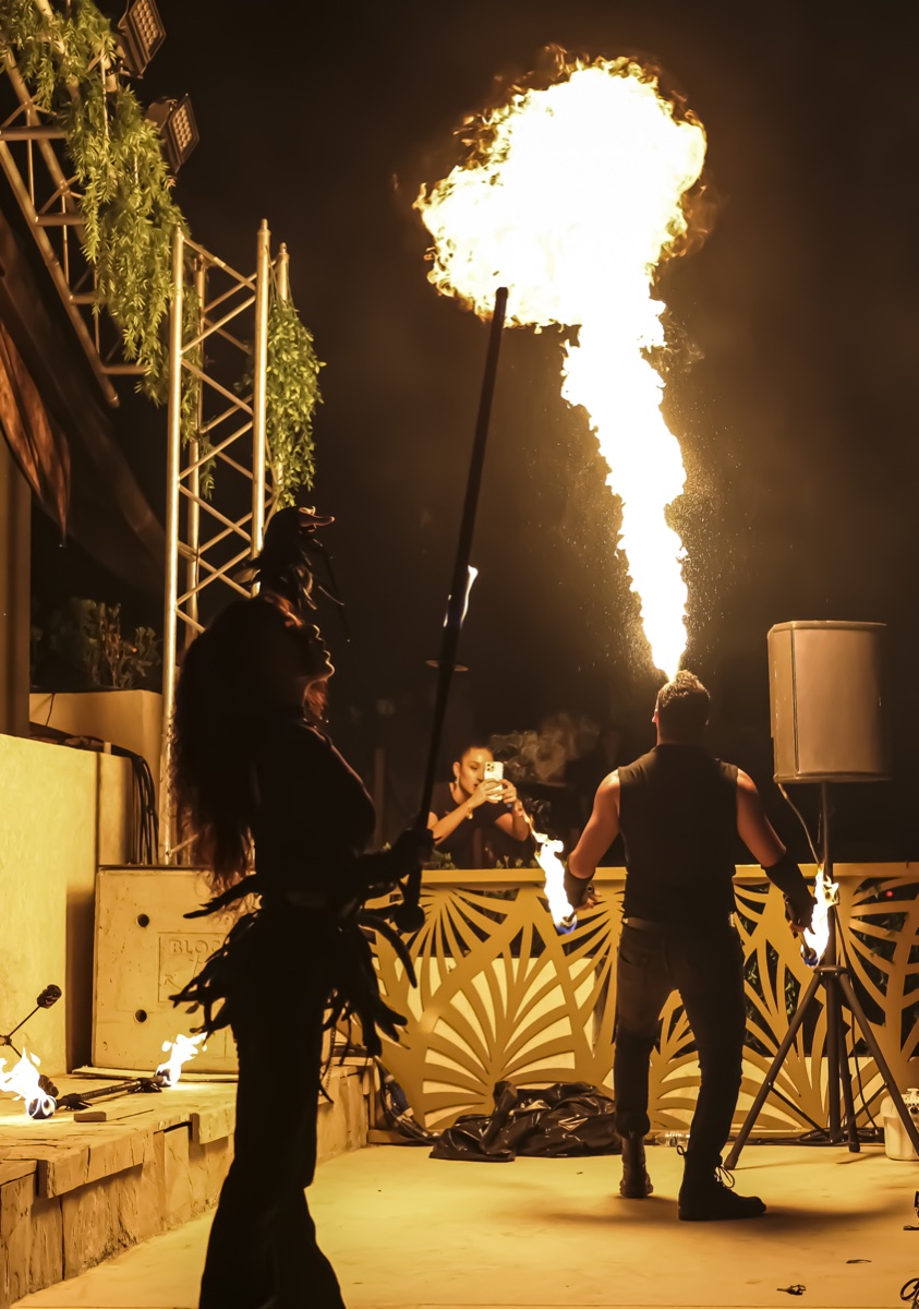 A fire-breather sending a flame plume skyward while a costumed performer stands silhouetted in the foreground