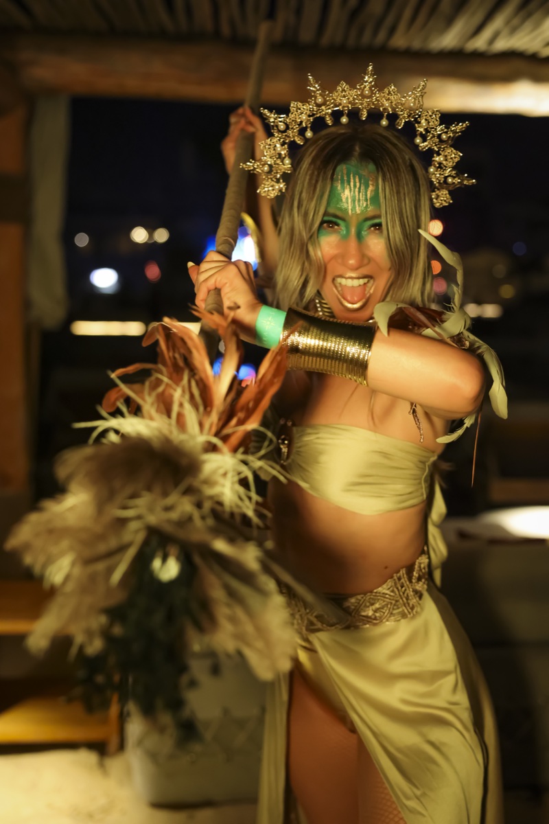 A performer in a gold crown and gold costume holding feather fans, laughing, at a night venue