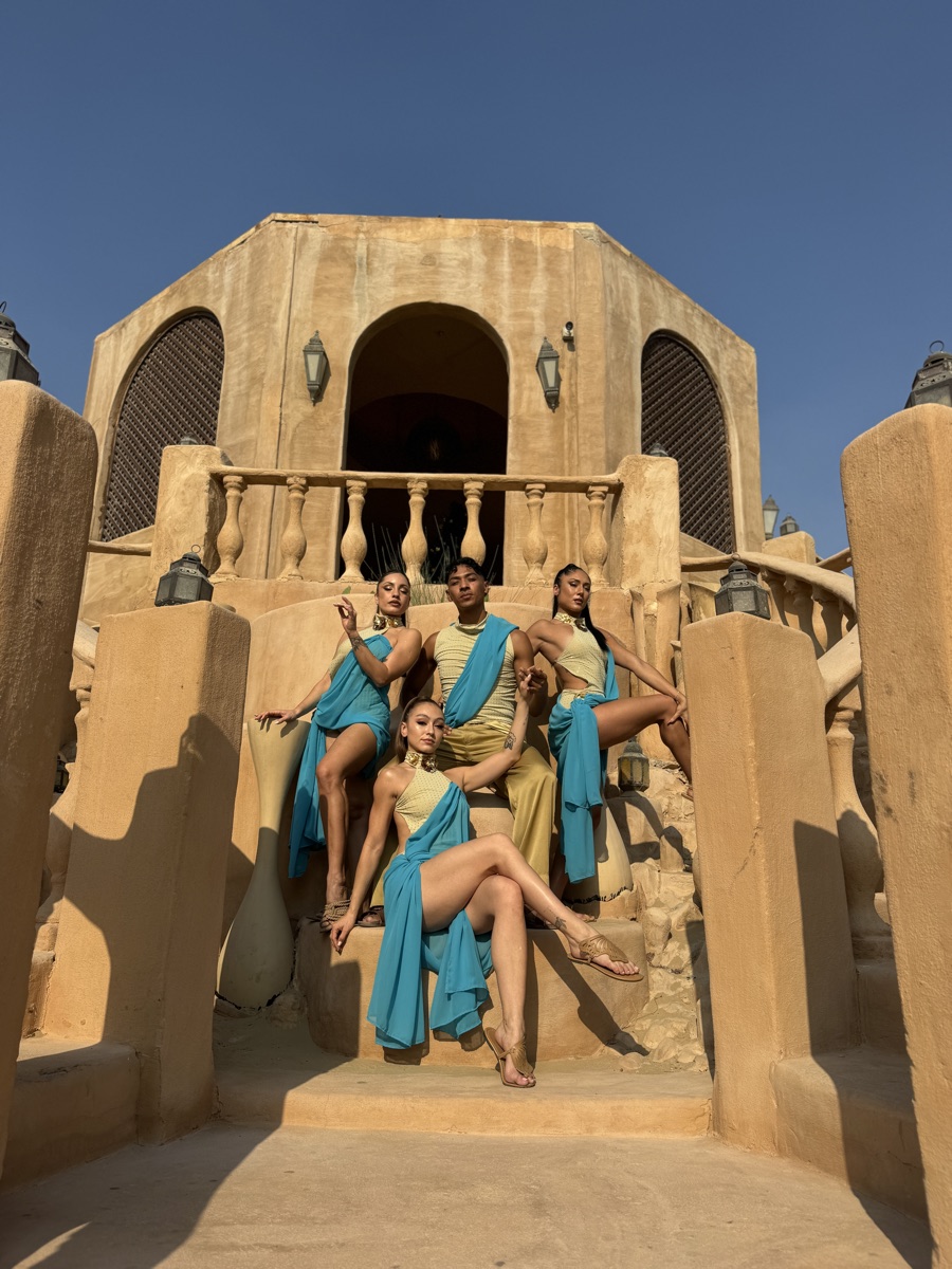 Four dancers in teal and gold draped costumes posed on a stone staircase