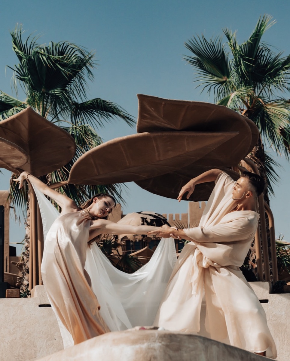 Two dancers in flowing ivory costumes stretching a length of silk between them beneath palms