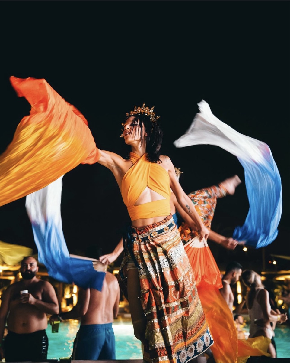 A crowned dancer swirling orange and blue silk veils at the poolside at night