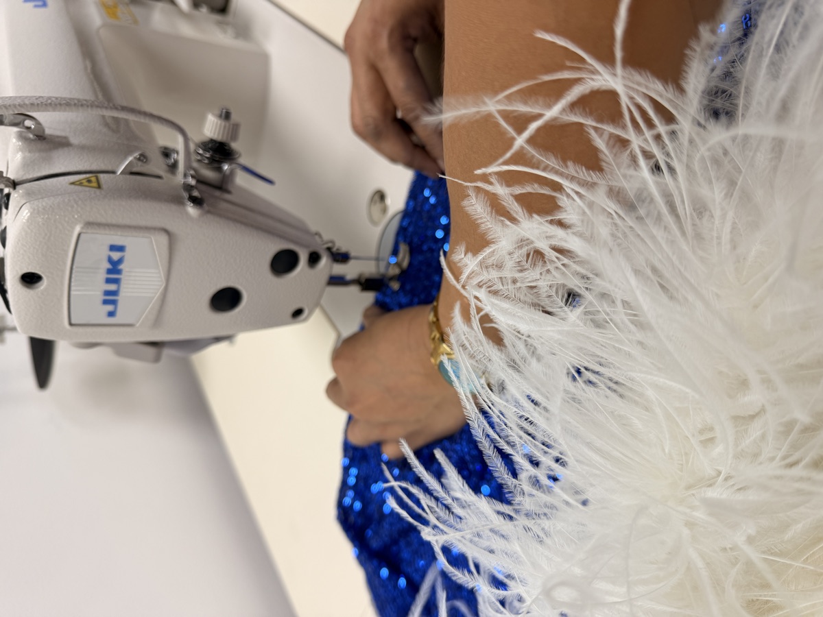 Hands guiding blue sequin fabric through a sewing machine beside white feathers