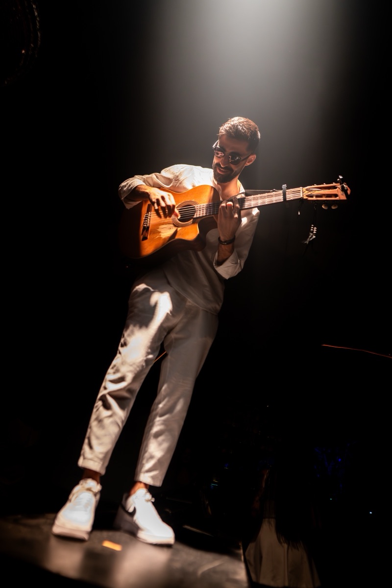 A guitarist in cream playing under a single spotlight on a dark stage