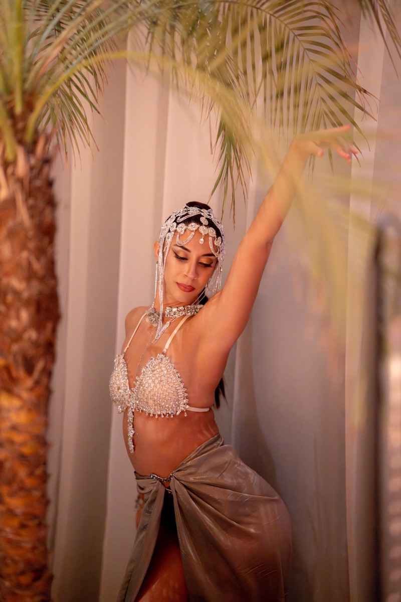 A dancer in a pearl and crystal costume posing between palm fronds in soft daylight