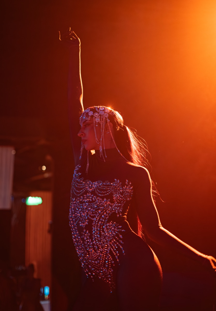A dancer in a crystal bodysuit rim-lit in amber haze, one arm raised