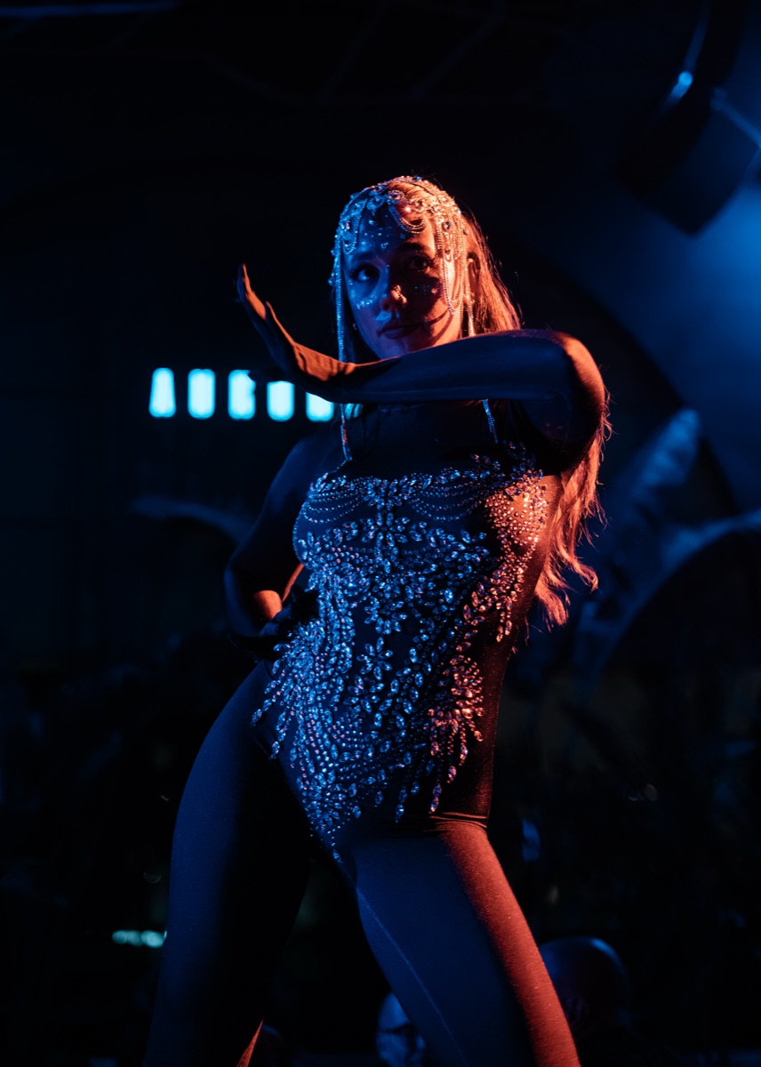 A dancer in a blue crystal bodysuit and long gloves mid-pose under cool blue club light