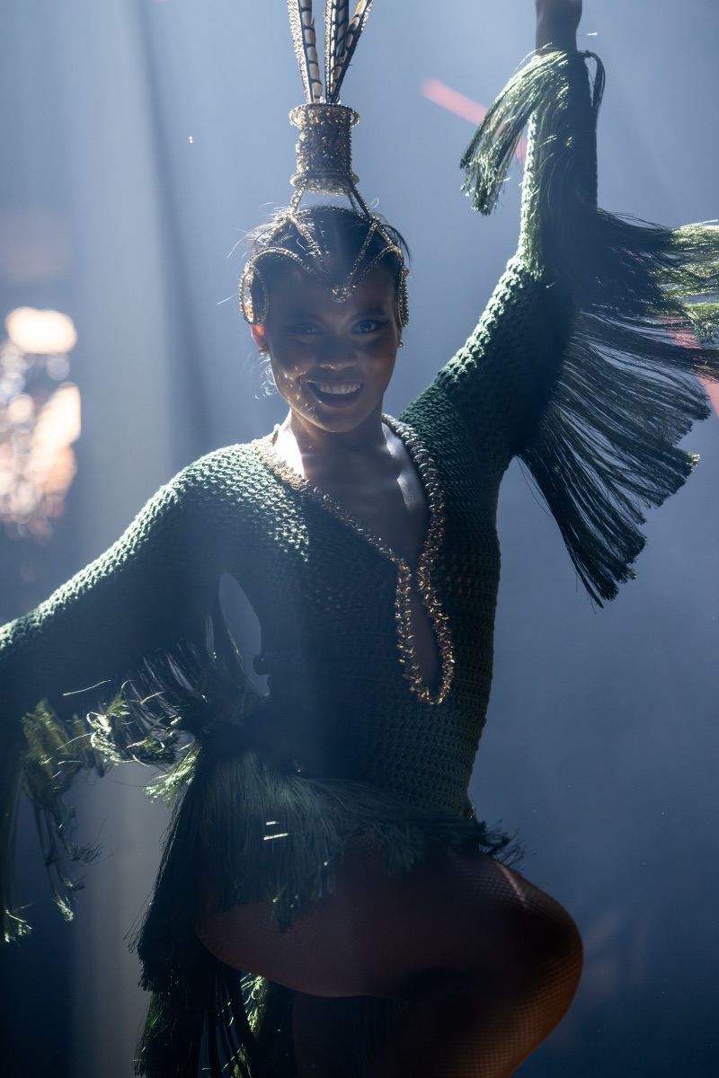 A performer in a green fringe costume and tall feather headdress wrapped in smoke