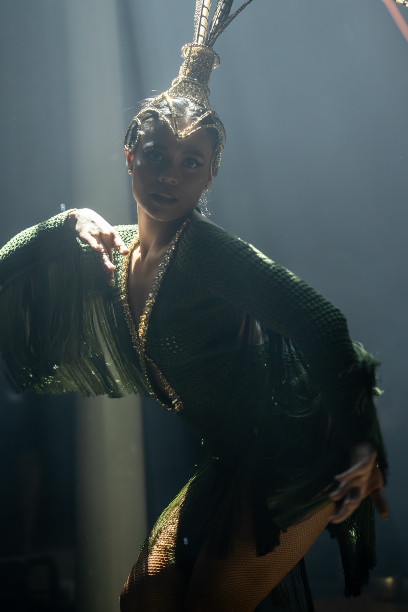 A performer in a green fringe costume and feather headdress, side-lit in smoke