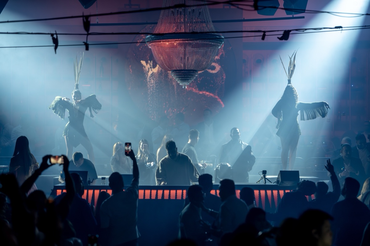 A wide production shot — feathered performers on podiums flanking a DJ booth beneath a chandelier