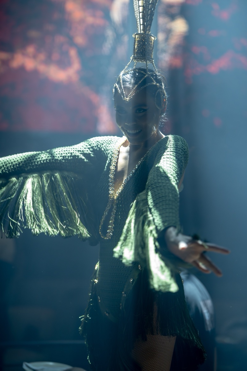 A performer in a green fringed costume with a mirrored headdress reaching through the haze