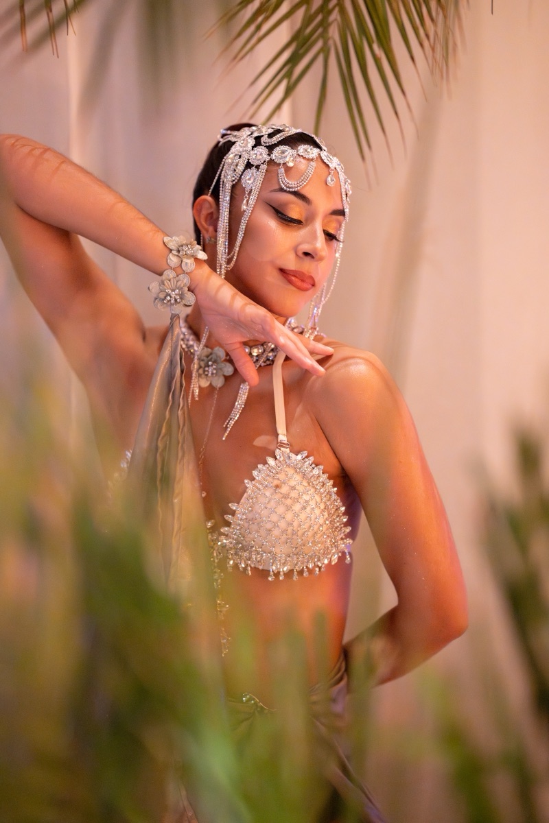 A dancer in a pearl-beaded costume framed by palm leaves against a warm cream wall