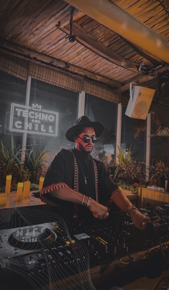 A DJ in a wide-brim hat at the decks beneath a neon sign