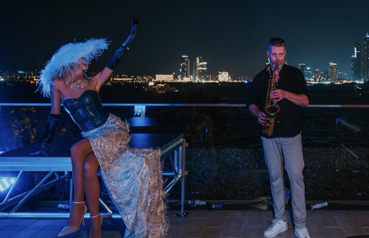 A saxophonist and a showgirl in a feather headdress performing on a rooftop above the Dubai skyline