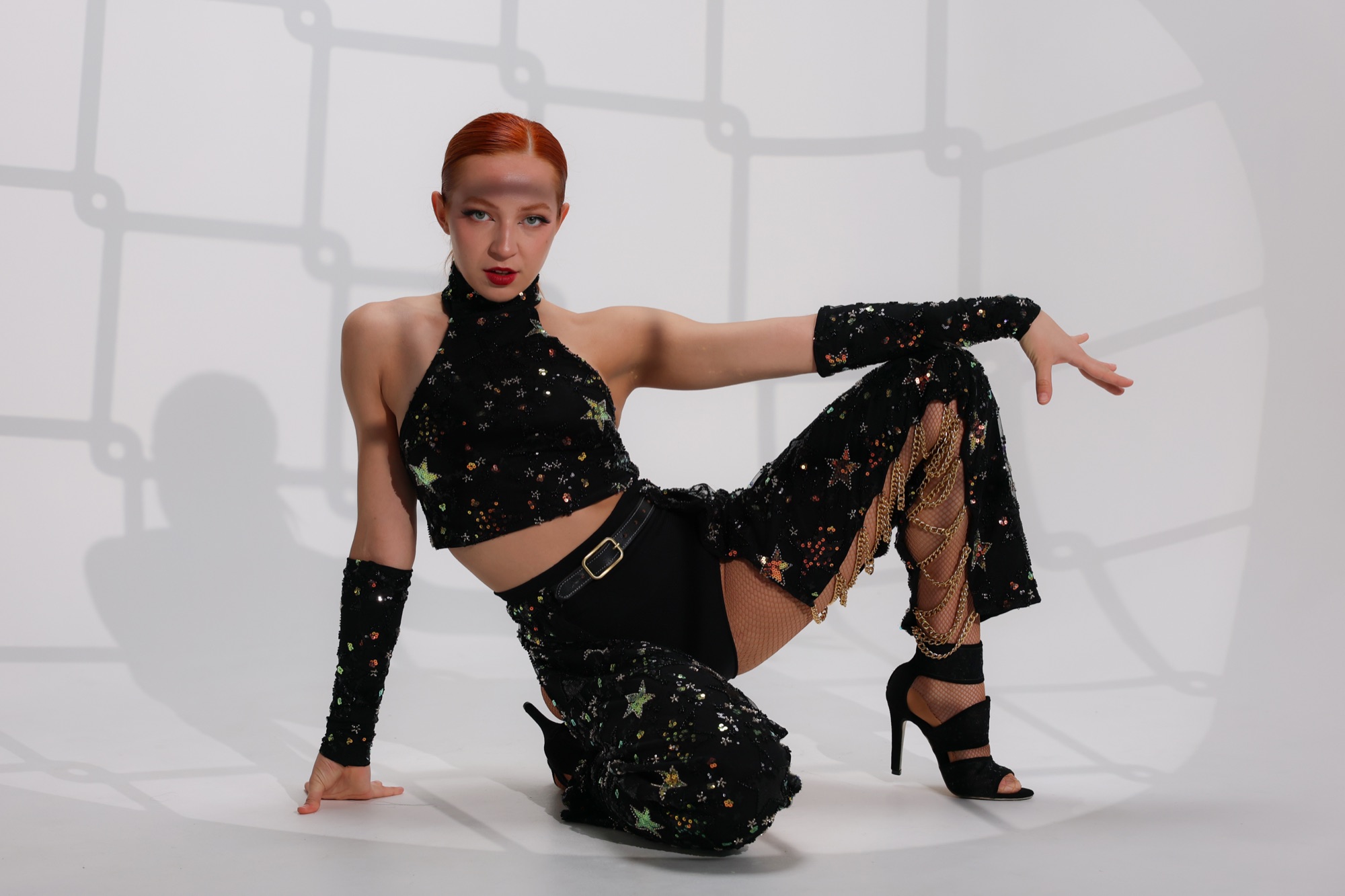 A Refined dancer in a black star-sequinned costume, seated on a pale studio floor in warm patterned light