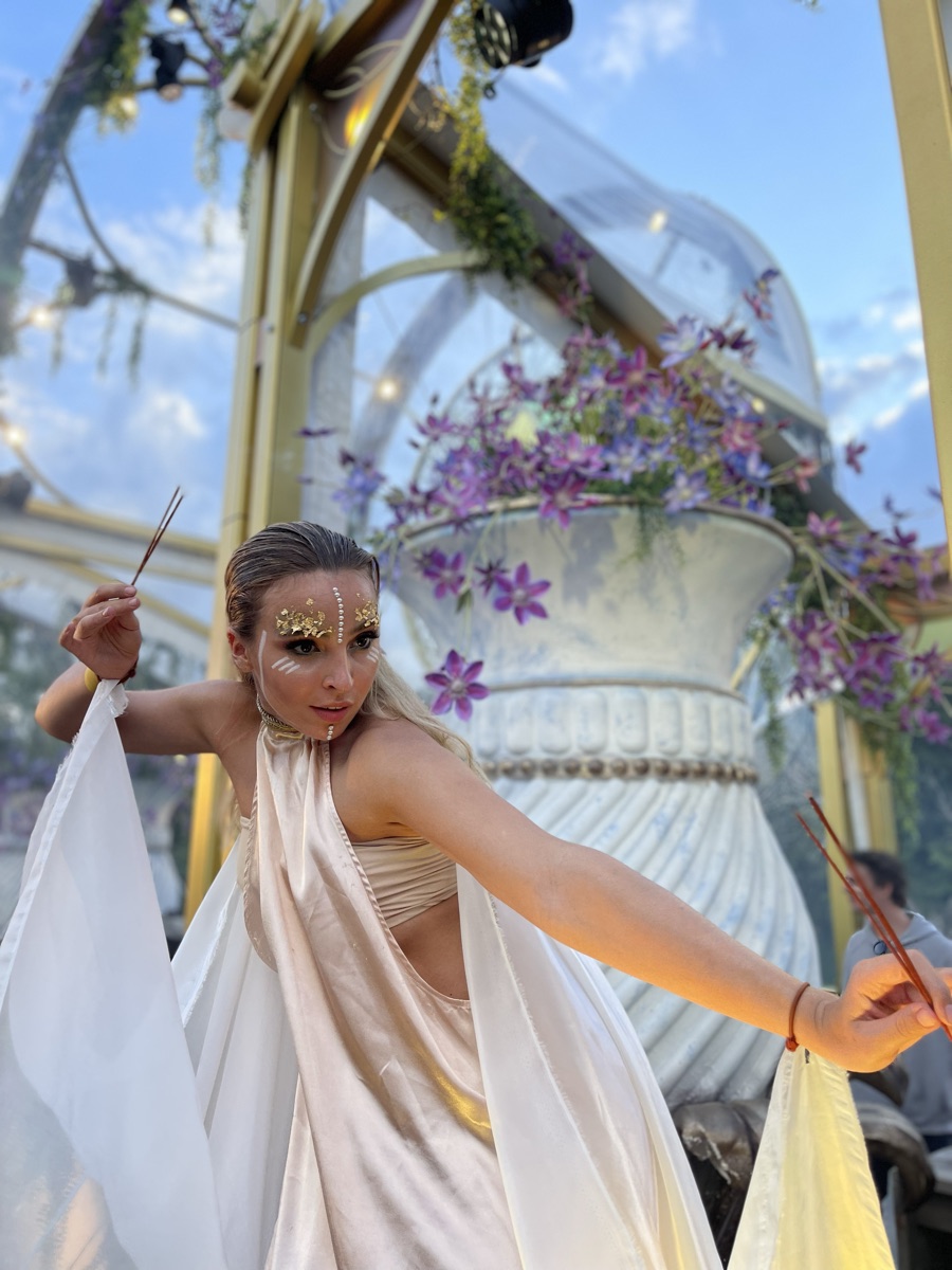 A performer in a white and gold draped costume with fans, posing under a floral arch in daylight
