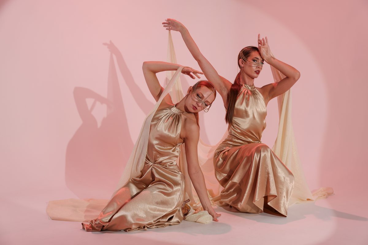 Two dancers in gold satin dresses posing against a pink studio backdrop