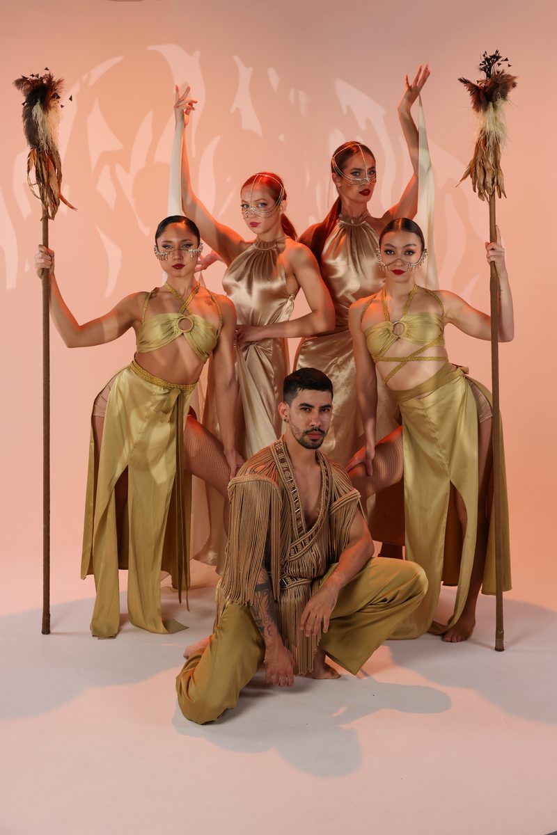 A Refined cast in gold costumes posed with tall staffs under warm light