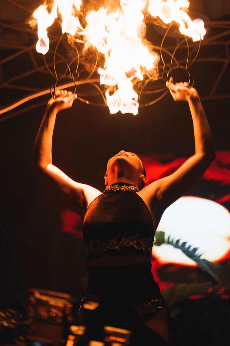A fire performer holding twin flaming fans overhead against darkness