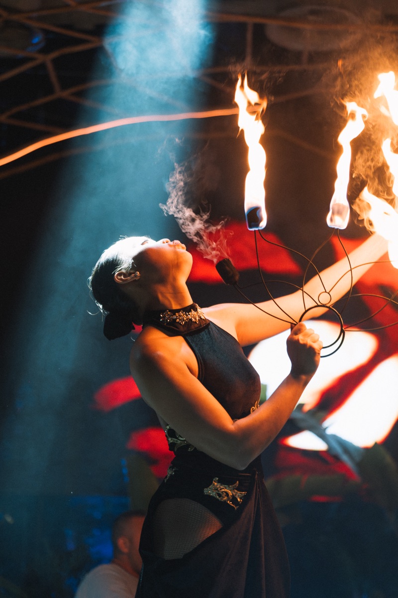 A fire performer exhaling smoke beneath raised torches, a blue beam behind