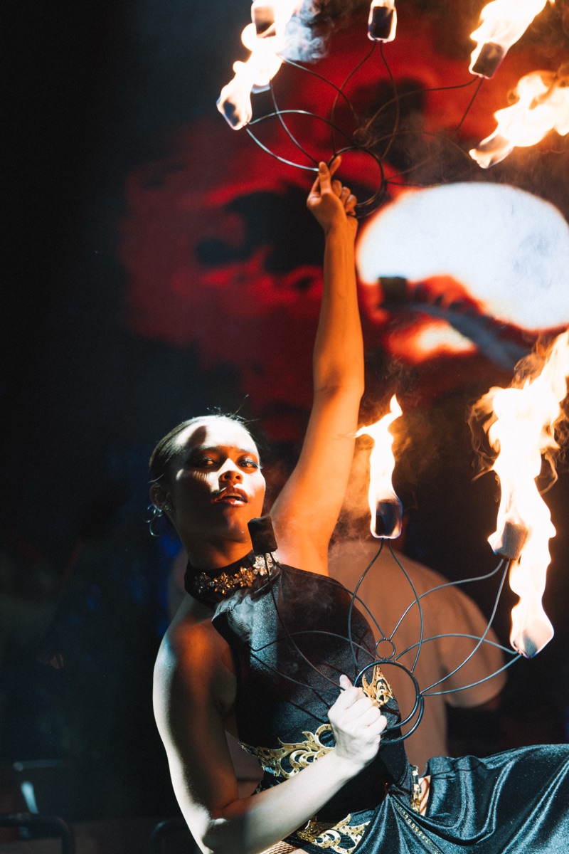A fire performer raising a burning fan against a red and black projected backdrop