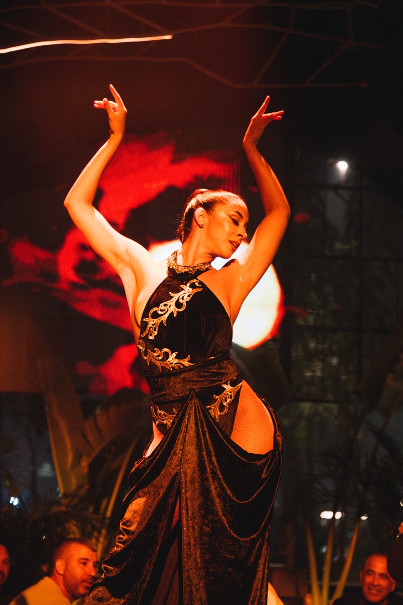 A dancer in a black and gold velvet gown with both arms raised, in front of a red-lit screen