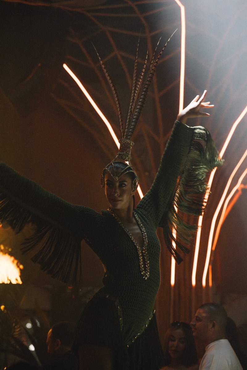 A performer in a green fringed costume with a tall pheasant-feather headdress, arms raised