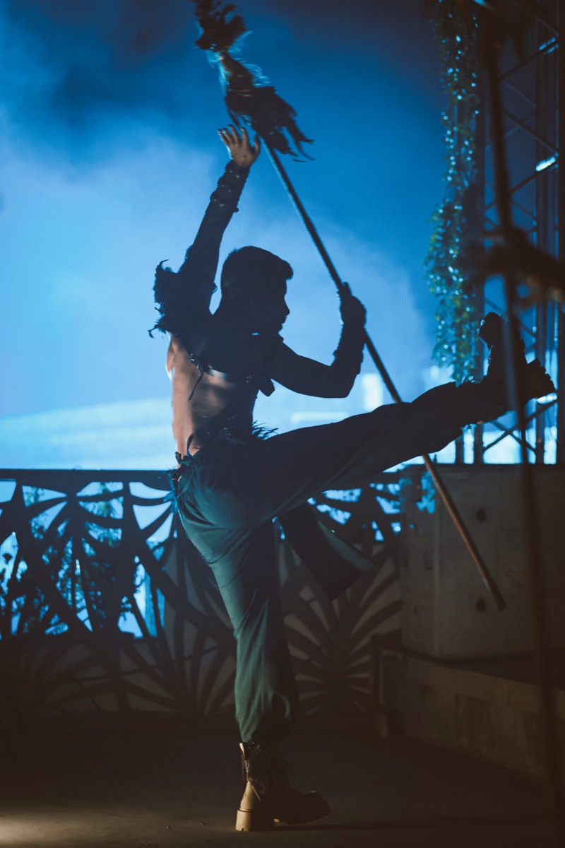 A silhouetted dancer high-kicking with a staff in smoky blue light