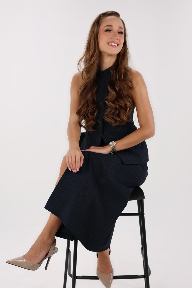 Laura, founder of Refined Performing Arts, seated on a stool in a navy pinstriped dress against a pale studio background