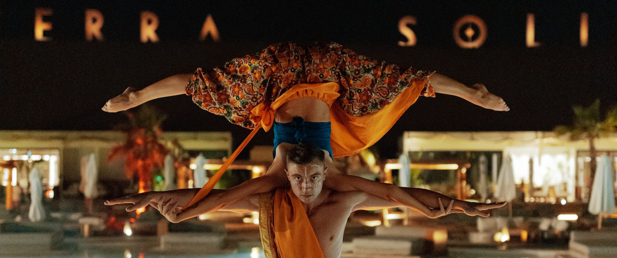 An acrobat holding a performer horizontally across his shoulders on a lit walkway between two pools at night, the Terra Solis sign glowing behind them