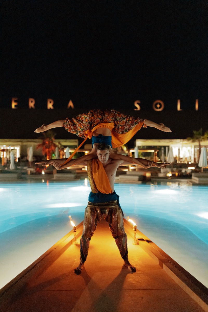 Two acrobats in a hand-to-hand balance on the walkway between the lit pools at Terra Solis